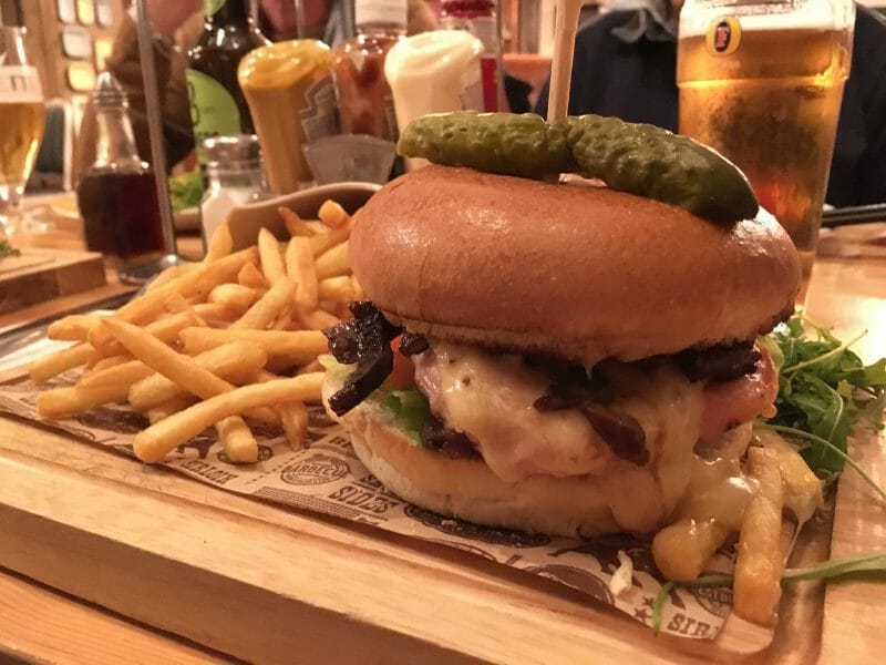 A chicken burger topped with lamb and cheese in a brioche bun, with a gherkin on top. Served with golden fries and rocket