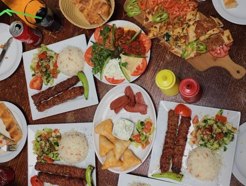 A flatlay of Turkish food including Adana kebabs, Turkish Pide (Turkish pizza), filo pastry triangles stuffed with feta cheese, sujuk, and more.