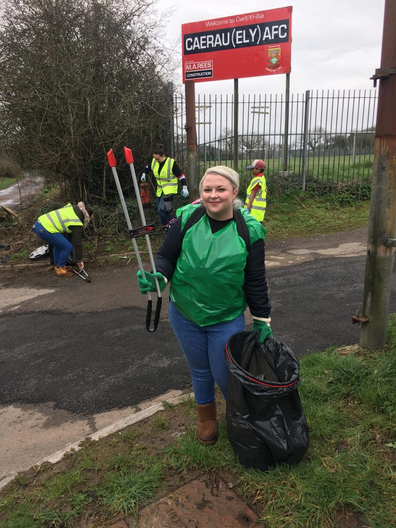 Me wearing a green plastic vest, holding a litter picker and black bin bag underneath the 'Caerau Ely F.C' sign
