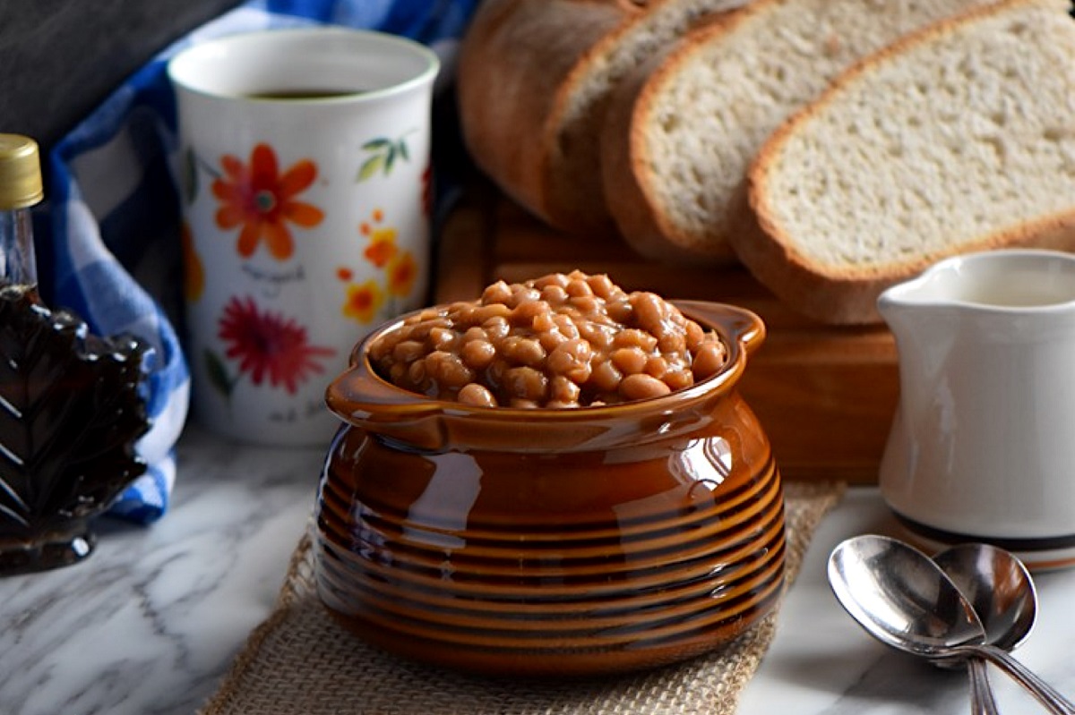 A pot of homemade baked beans, with a loaf of bread and a cup of coffee in the background.