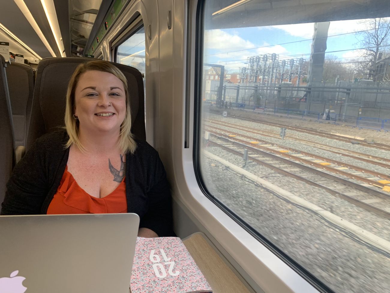 Girl in table seat on train, with laptop and diary, looking at the camera and smiling