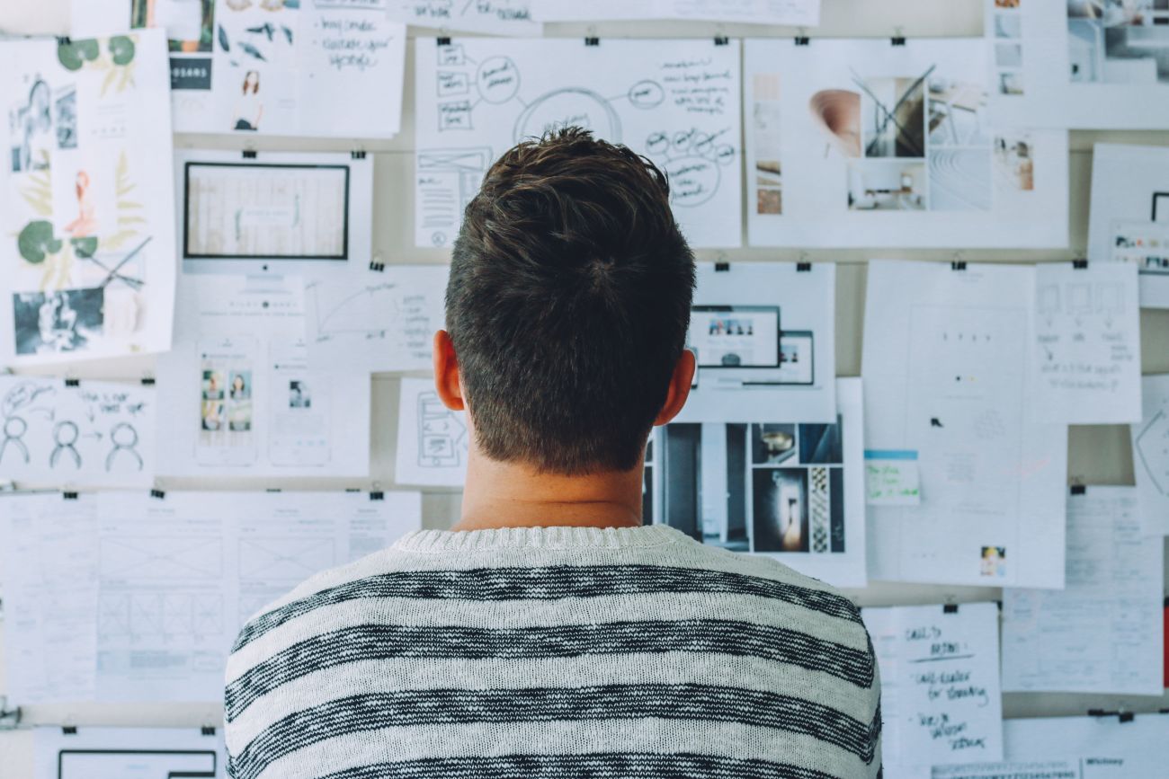 Man photographed from behind, staring at a wall covered with notes and plans. Having a plan B is an important part of preparing for your next big trip.