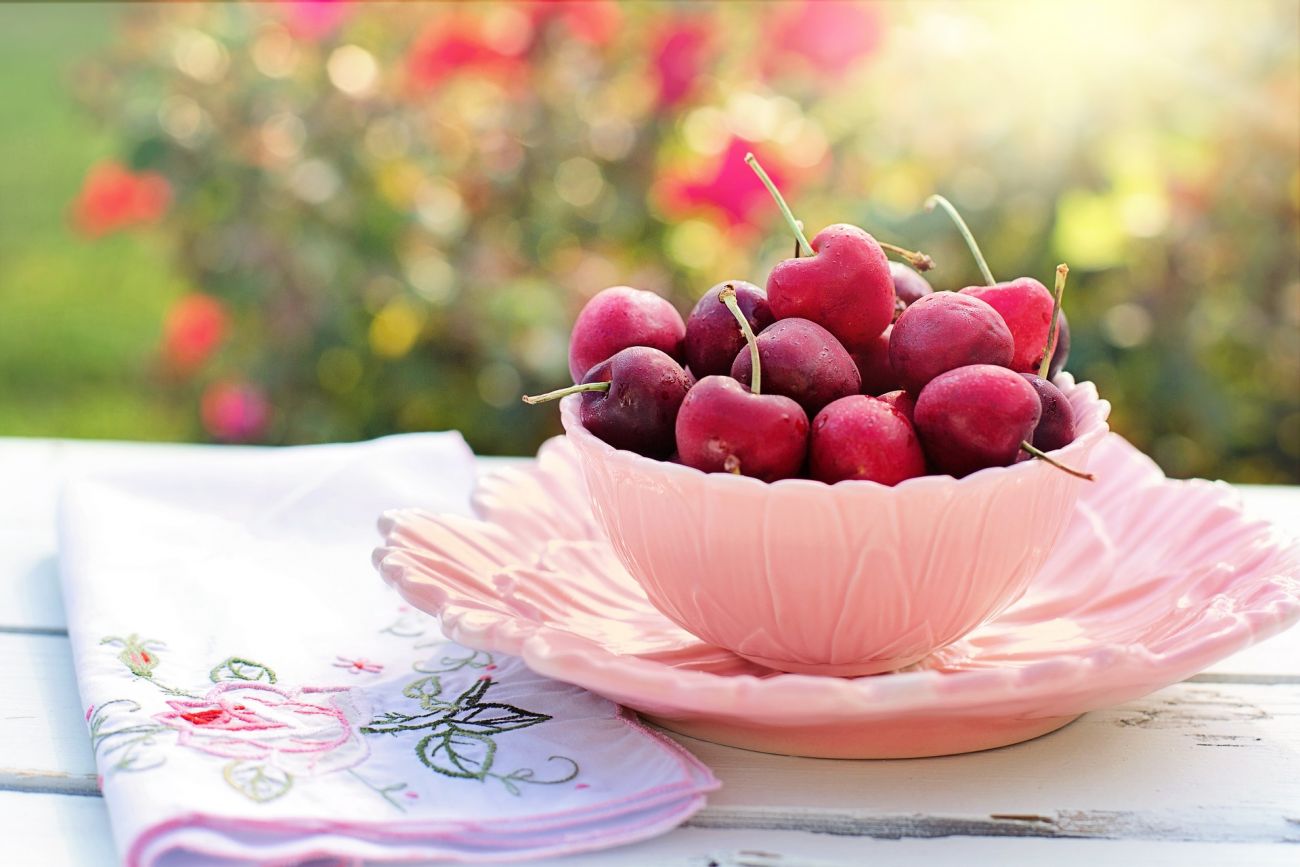 A bowl of fresh red cherries sitting on a saucer next to a tea towel on a garden table.