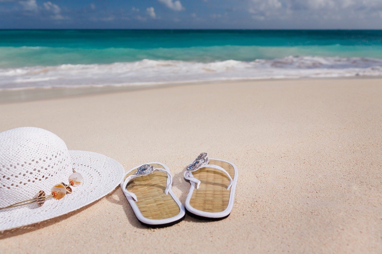 A pair of jewelled flip flops sitting on a tropical beach next to a white straw sun hat