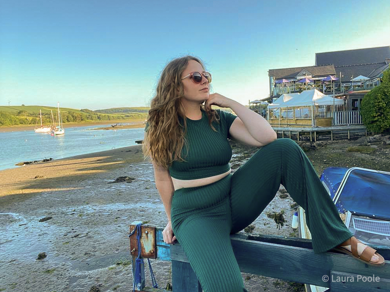 A lady wearing an emerald green co-ordinating set in a coastal scene, sitting on a fence.