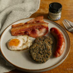 Welsh Lavercakes, made using Welsh Laverbread, on a traditional Welsh breakfast with fried egg, sausage and Welsh back bacon, with a jar of laverbread and cutlery in the background.
