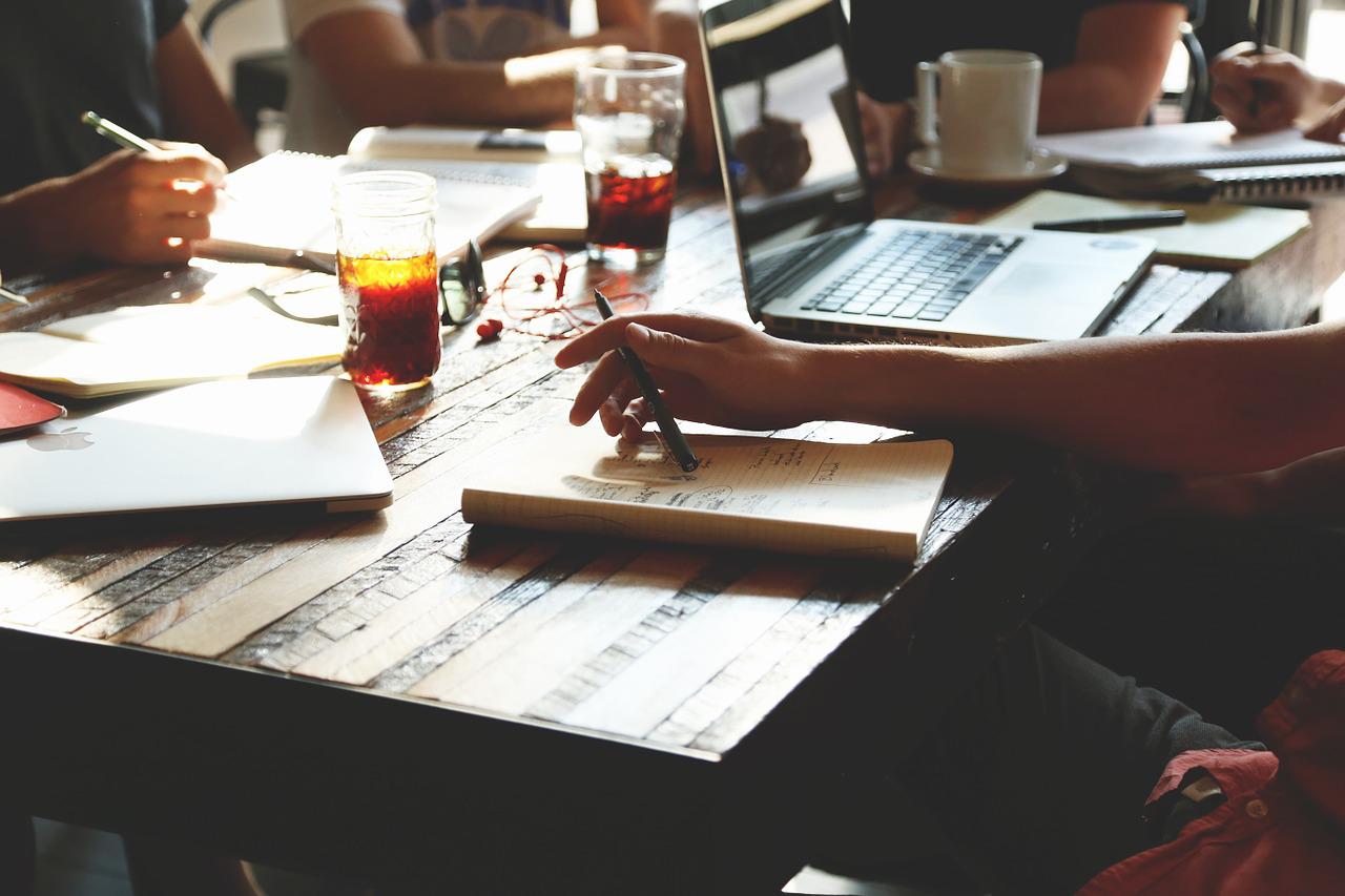 A group of people gathered around a table with laptops, notebooks and pens.