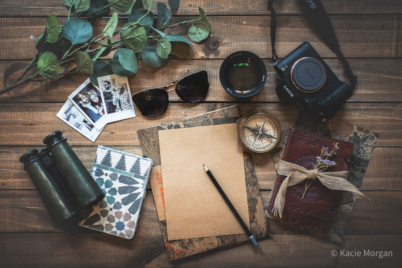 A selection of travel accessories laid out on a table - binoculars, photos, notebook, pencil, sunglasses, passport, camera and compass.