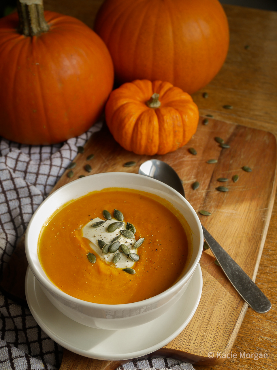 Pumpkin coconut and chilli soup served in a bowl on a chopping board, scattered with pumpkin seeds and surrounded by a tea towel, two large pumpkins and a smaller munchkin pumpkin