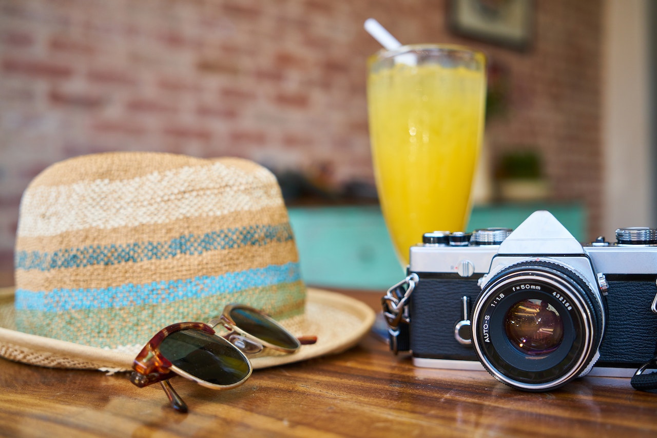 Sunglasses, a straw sun hat and a camera, next to a glass of orange juice