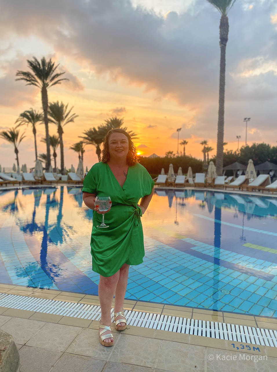 Me standing next to the outdoor pool at Asimina Suites Hotel in a green satin dress, drinking a glass of gin.