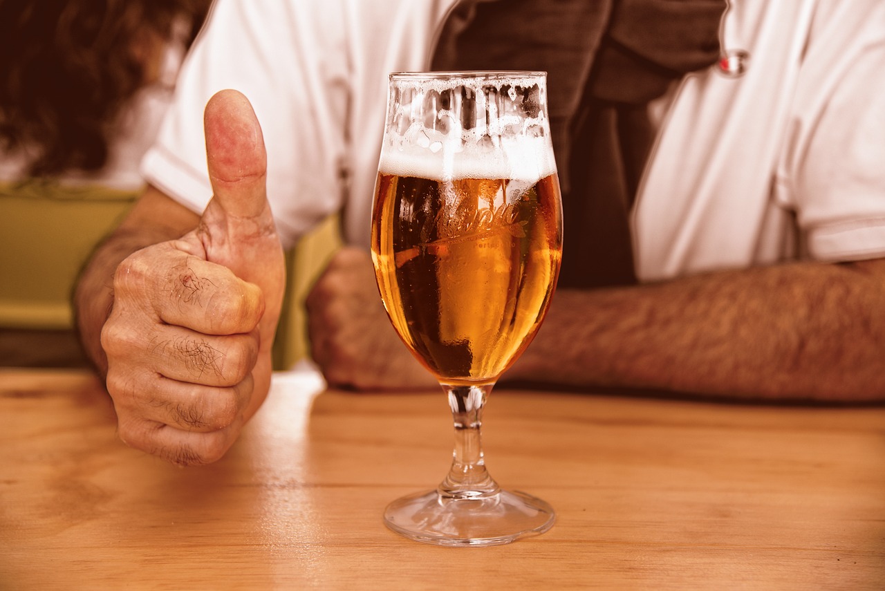 A glass of beer on a wooden bar, with a man holding his thumbs up next to it.