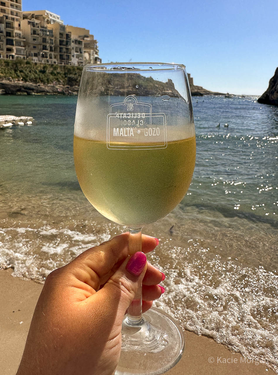 A wine glass filled with white wine, bearing the words 'Malta, Gozo' pictured against a backdrop of a beach and the shimmering sea.