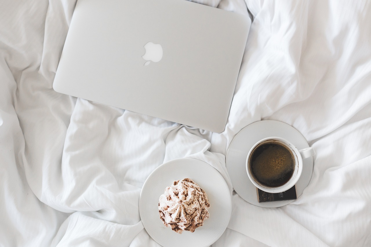 A coffee, muffin and laptop sitting on a crisp white bed.