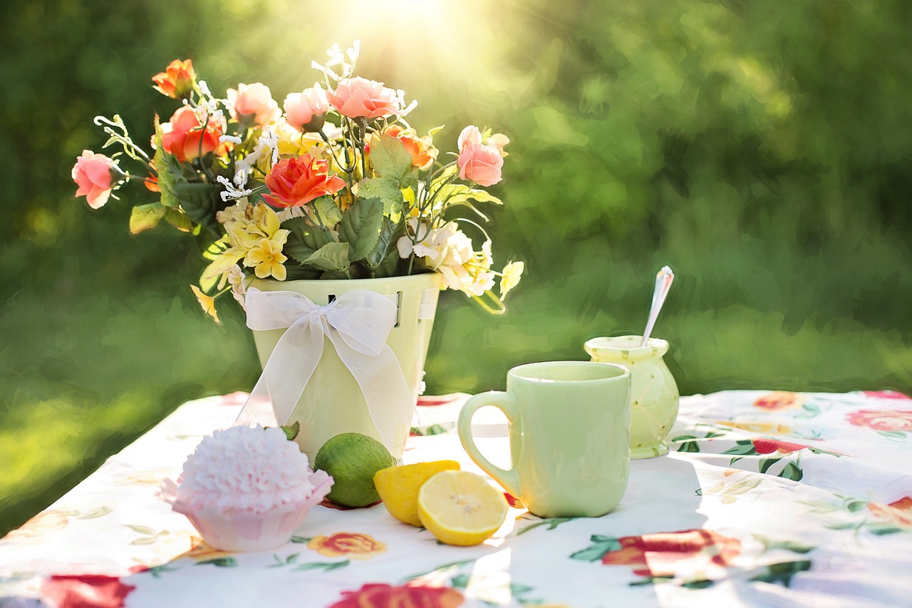 A vase of flowers, a cup, sliced lemon and lime, and a cupcake, on a floral tablecloth in a garden.