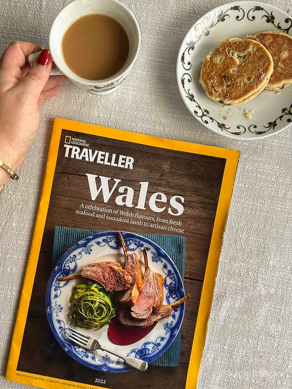 National Geographic Traveller UK's November 2023 supplement on Welsh food and drink. Next to it, there is a hand holding a cup of tea, and a sauce containing two Welsh cakes.