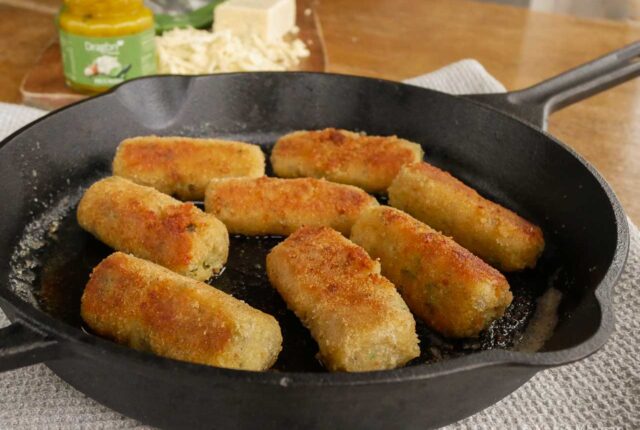 Glamorgan sausages in a cast iron frying pan, placed on top of a teatowel on a wooden table. In the background, you can see Dragon Piccalilli Pickle and grated Dragon cheese on a chopping board.