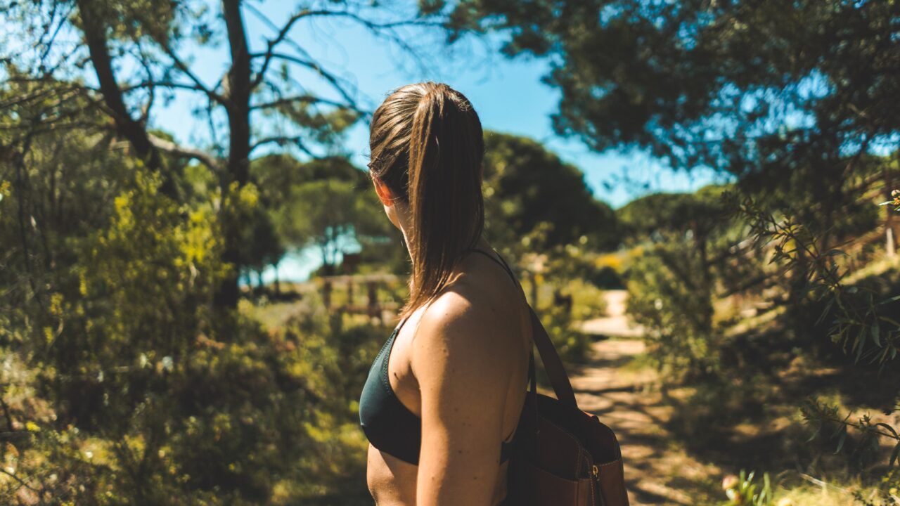 A girl walking down a path through the countryside wearing a black crop top bra, with her hair in her ponytail. She is looking backwards in the direction she came from.