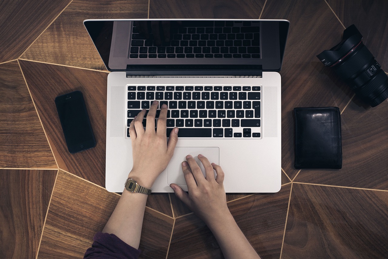 Hands typing on a Macbook on a wooden table, with a black phone and a wallet either side.