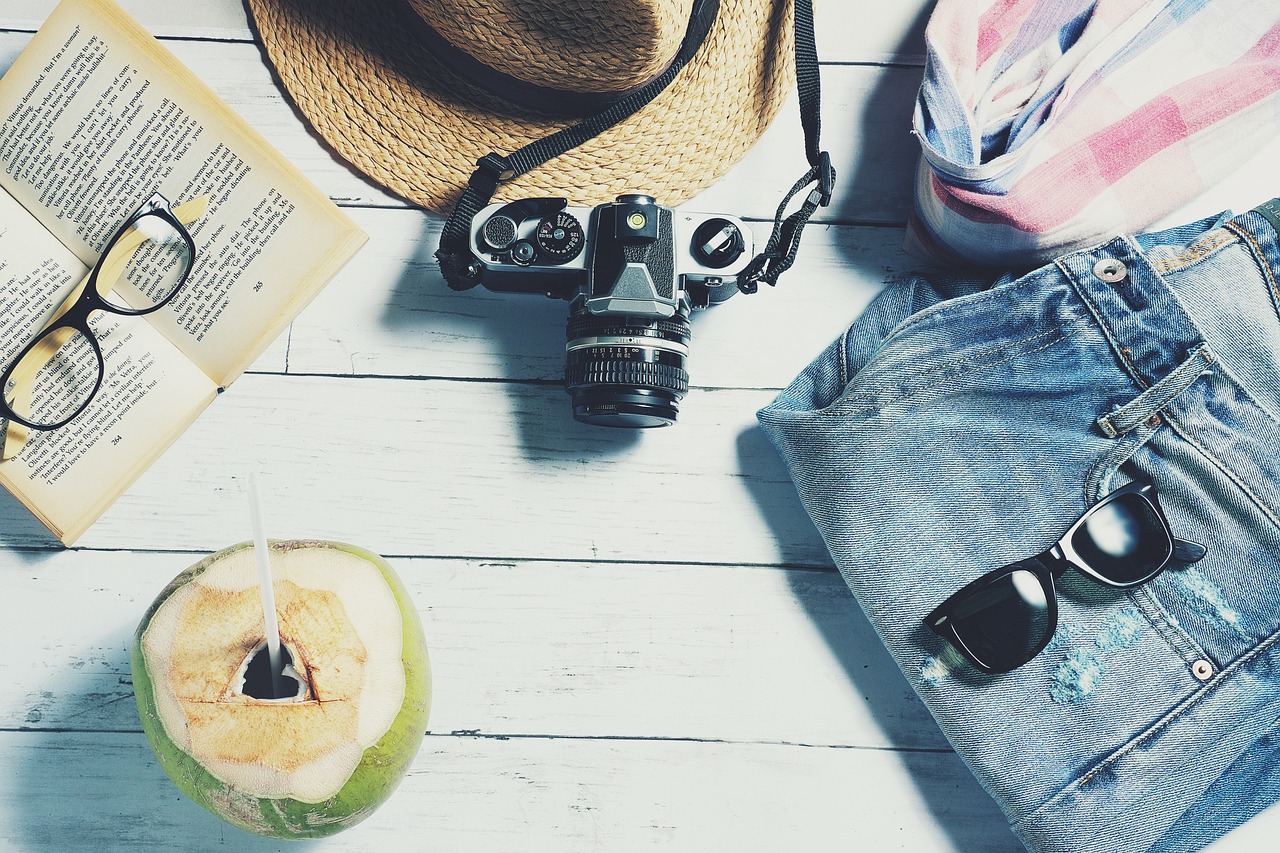 A book with glasses on top of the pages, a camera, a pair of jeans, sunglasses, a fresh coconut with a straw in it, a hat, and a scarf.