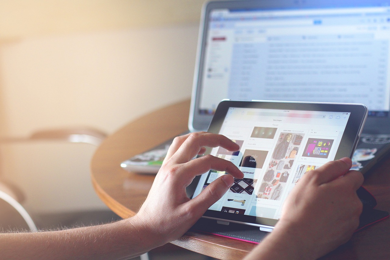 Hands/fingers touching images on a tablet, with a computer screen in the background