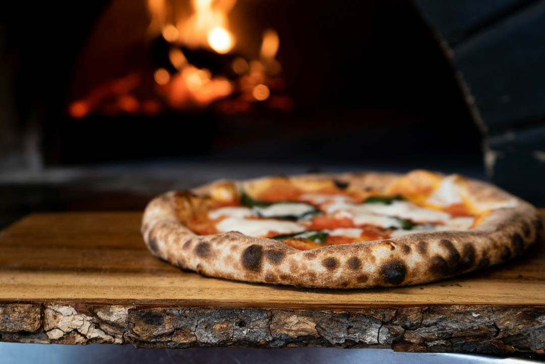 A pizza, cooked, next to a wood-fired Italian pizza oven.