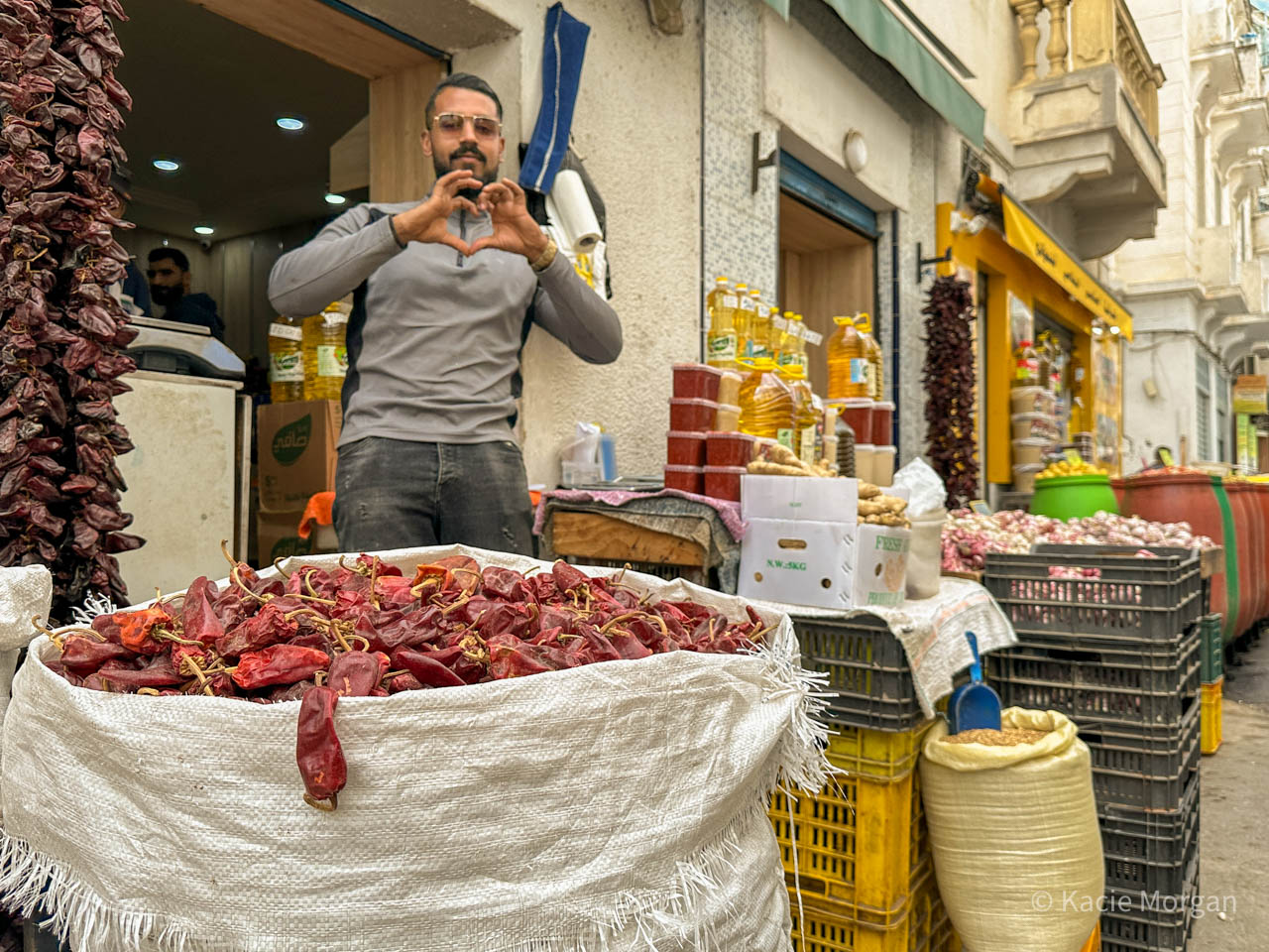 A man selling chillies on a stall. He is making a heart-shaped gesture at me with his hands.