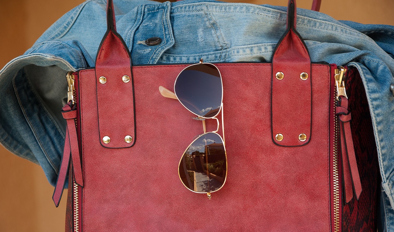 A pair of sunglasses hooked onto a red leather bag, with a denim jacket placed through the handle.