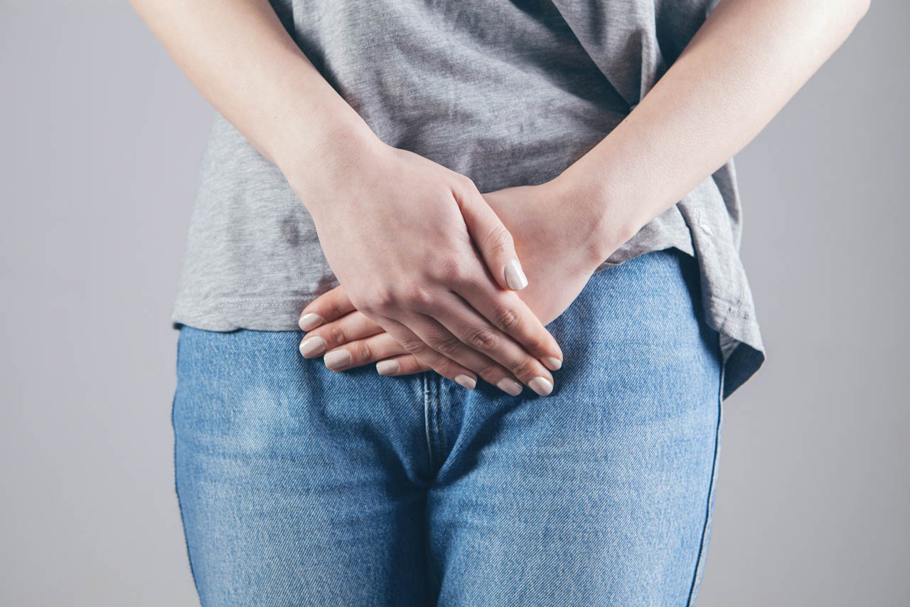 A lady wearing jeans and a grey tshirt, photographed close-up (her face isn't visible) with her hands folded over her abdominal area.