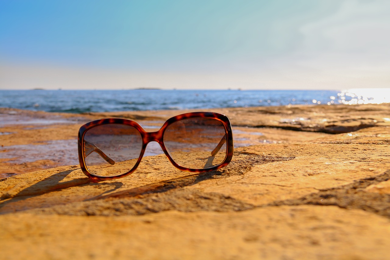 A pair of sunglasses on a rocky beach, with the sea visible behind.