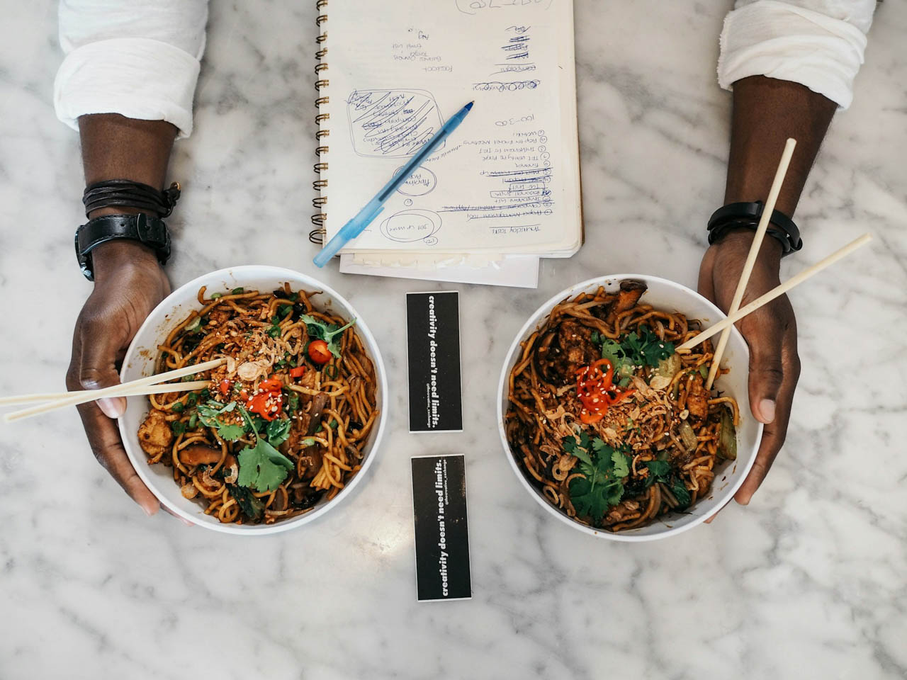 Two hands holding bowls of Thai noodles, topped with red chillies