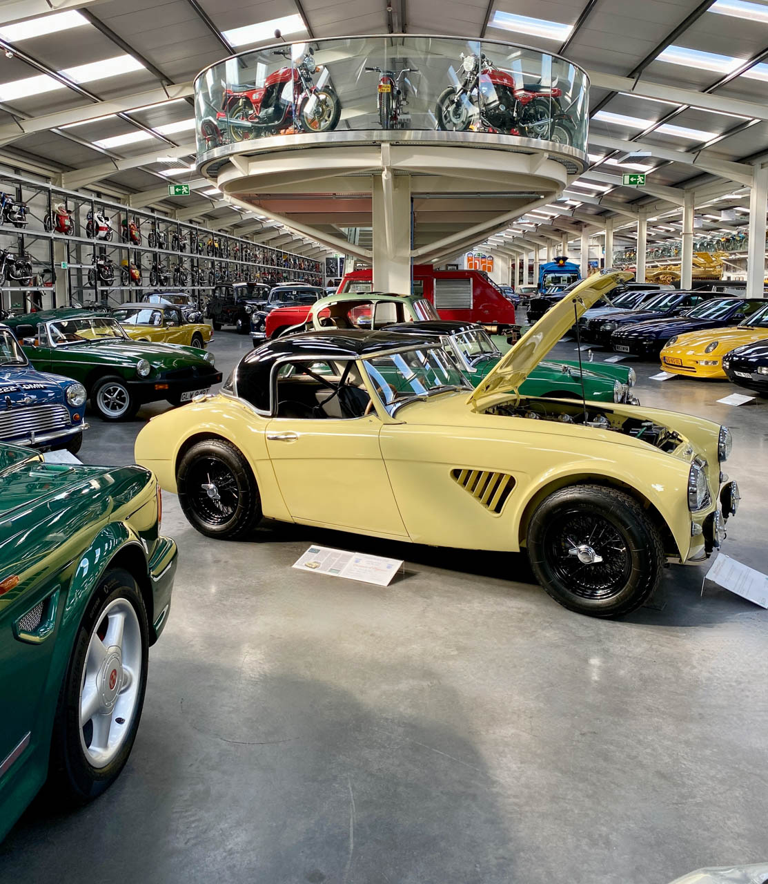 Vintage cars and motorcycles at a motor exhibition. A pale yellow car appears upfront.