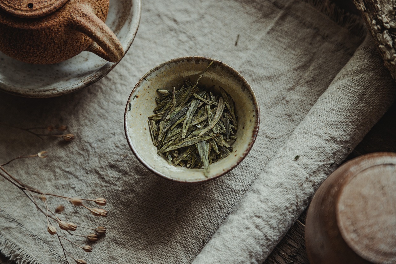 A cup of green tea leaves next to a teapot, on a linen tablecloth