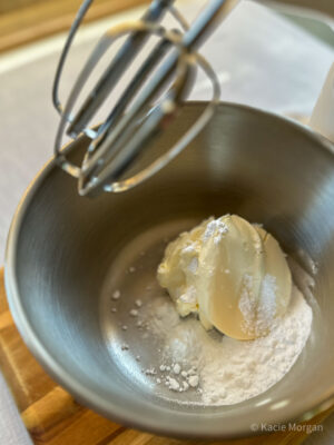 Creams cheese, icing sugar and vanilla essence in a mixing bowl, with the mixers overhead