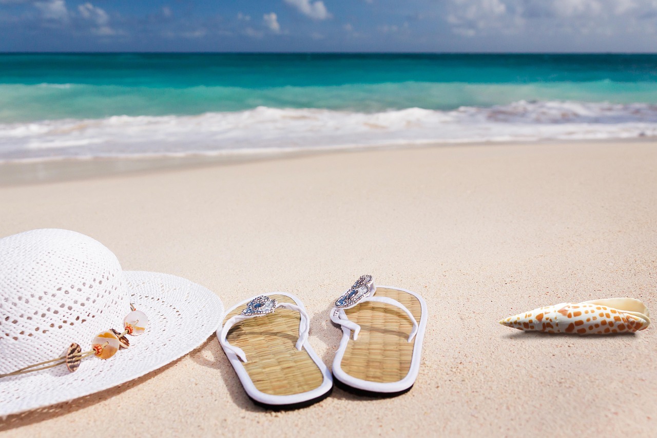A sun hat, embellished flip flops and a shell on a beach, with the sea visible in the distance