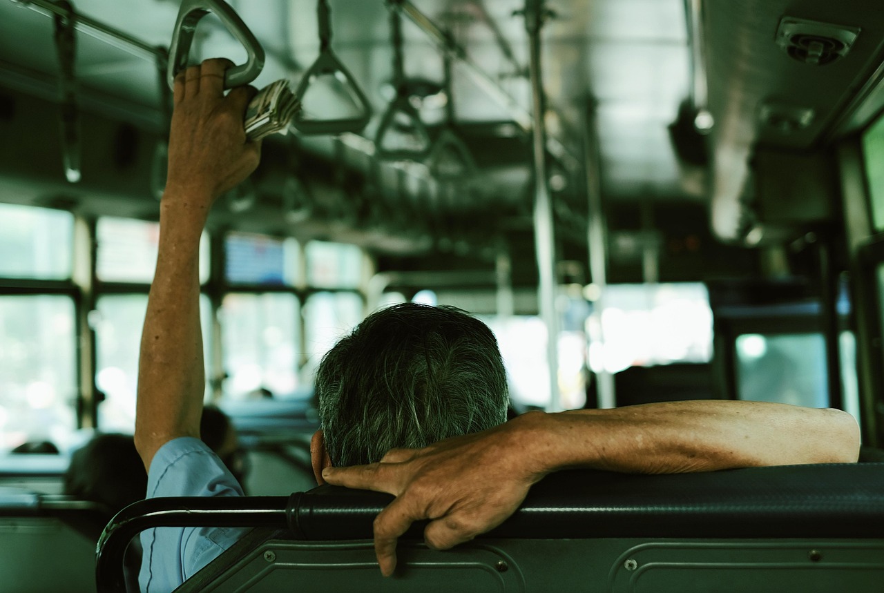 A man sitting on a bus holding the hand rail of the seat behind him with his right hand, and holding the overhead hanging handles with his left hand.