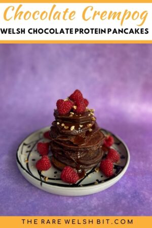 A stack of chocolate crempogau (Welsh pancakes) topped with chocolate sauce and rasperries on a plate.
