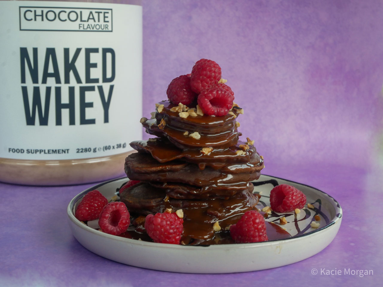 Chocolate protein pancake stack topped with raspberries, chopped hazelnuts and chocolate sauce, with Naked Whey protein powder behind