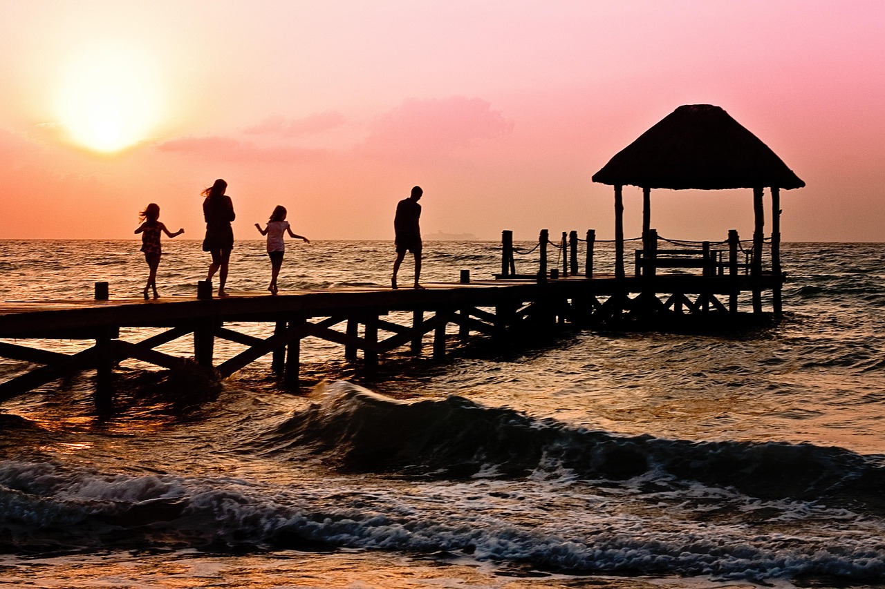 The silhouette of a family walking across a wooden board walk to reach a small hut sitting on the sea.