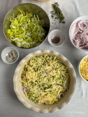 Assembling the Welsh onion cake by creating layers of sliced potato, sliced onion, finely sliced leeks, grated cheese and fresh thyme.