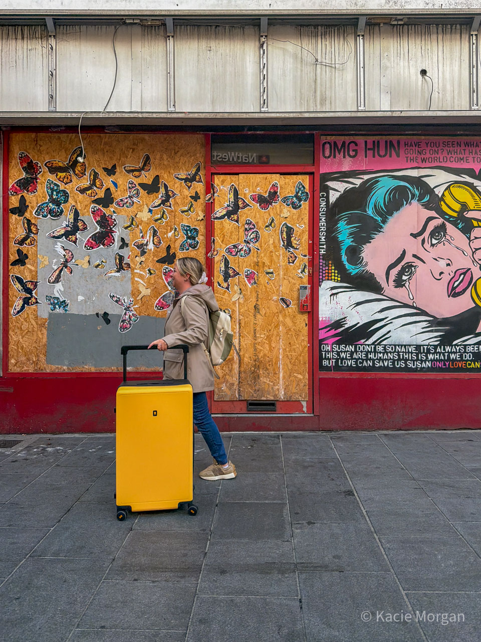 Kacie walking past a quirky looking shop doorway in Newport, south Wales with her yellow LEVEL8 Voyageur suitcase