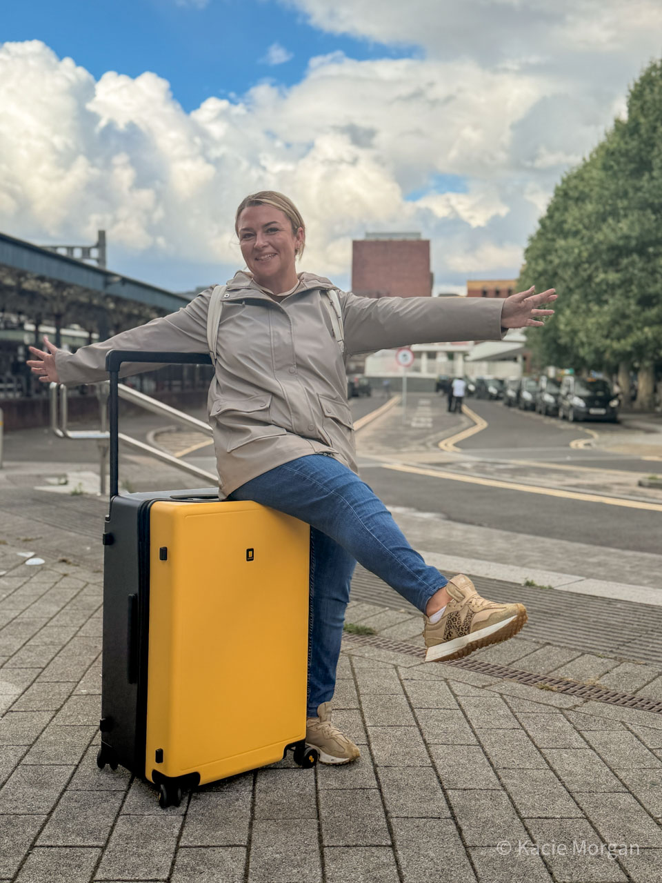 Kacie sitting on her suitcase with her arms stretched out, smiling.