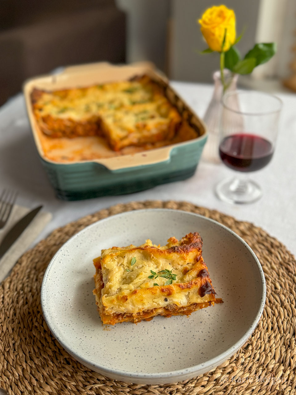 PGI Welsh Lamb lasagne with Caerphilly cheese bechamel sauce, served in a pasta dish with the full lasagne dish visible behind, next to a glass of red wine and a yellow rose.