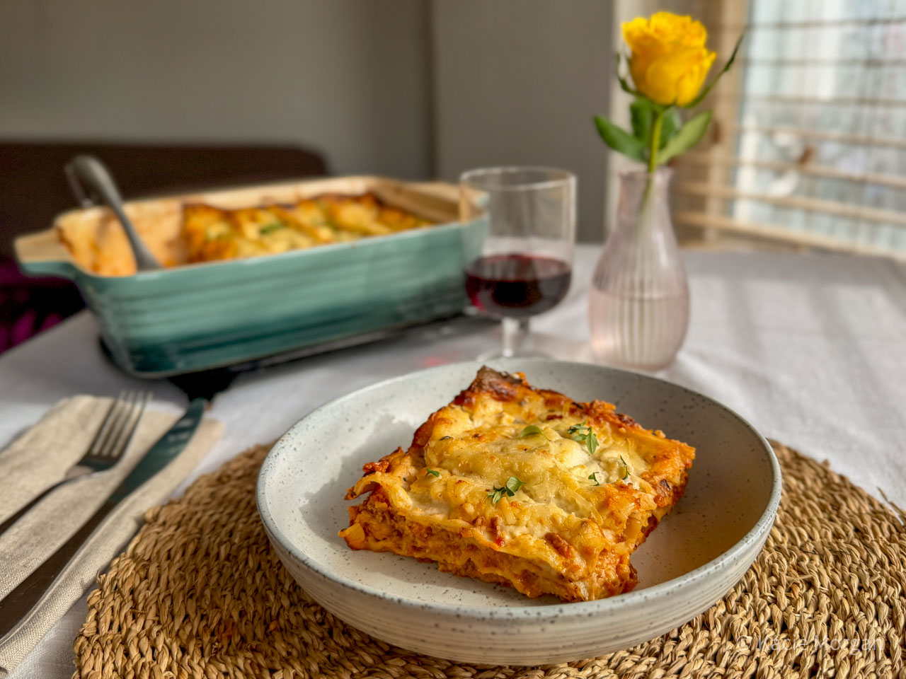 PGI Welsh Lamb lasagne with Caerphilly cheese bechamel sauce, served in a pasta dish with the full lasagne dish visible behind, next to a glass of red wine and a yellow rose.