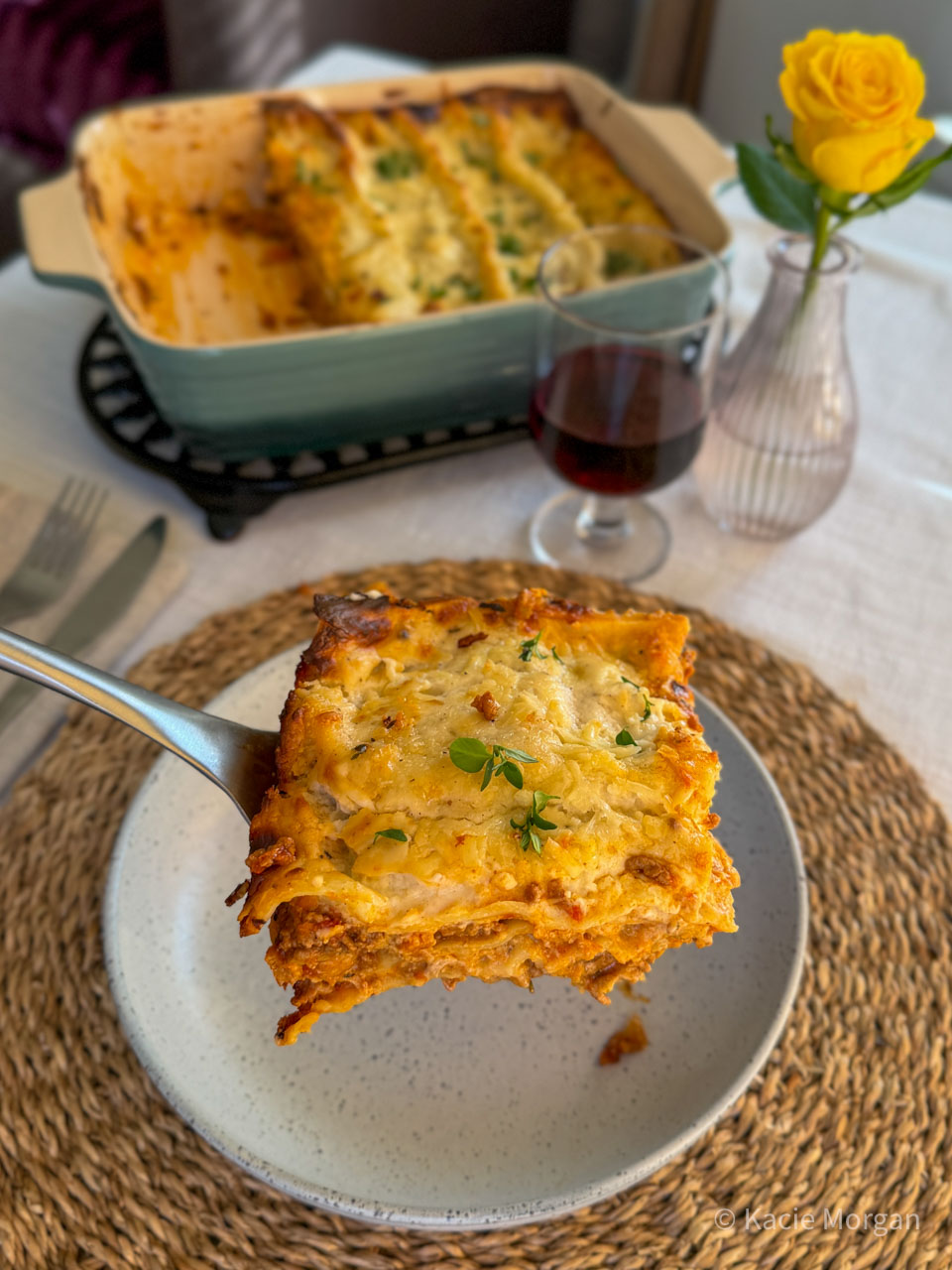 A portion of Welsh Lamb lasagne suspended on a flipper overhead a pasta bowl, with the full lasagne dish visible behind.