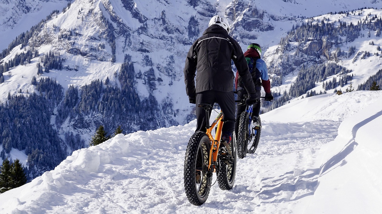Two cyclists on fatbikes riding a snowy alpine trail in winter mountains.